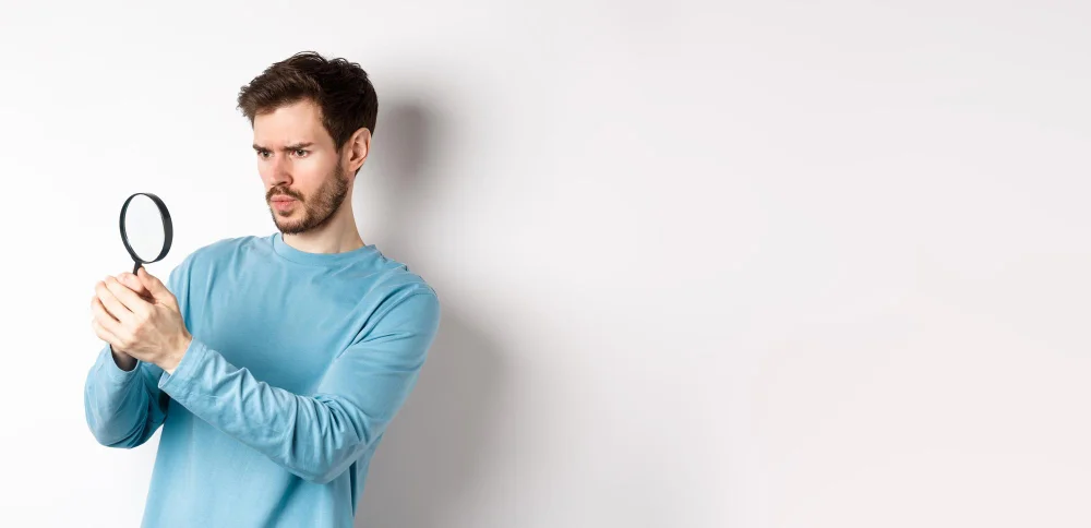 Man in blue sweater closely examining with a magnifying glass.