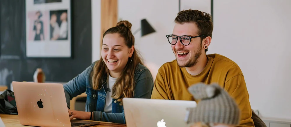 Two people sitting at a table, smiling while looking at their laptops.
